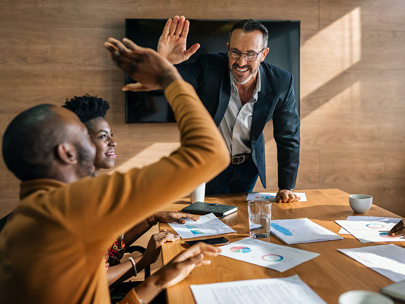 A seated, young man high-fives a man with glasses who is standing, in an office boardroom.